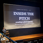 Anne Russell speaks at a podium in front of a large screen displaying “Florida Film Festival 2026 Inside the Pitch: creating a pitch deck live. Can a room of strangers build a film pitch in 30 minutes?”