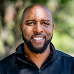 Full Sail grad Sidney Walker in the foreground smiling while wearing a black windbreaker with lush greenery behind him.