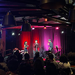 Three performers sing on a stage in front of an audience. Red and pink lights shine behind them.