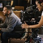 Behind the scenes on a student film set, one student sits in a chair with a notebook while another student works behind a film camera