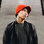 A young Asian man wearing a red knit cap, black jacket and shirt, green pants, and a burgundy bag stands between two escalators. His hands are in his pockets and he is looking off to the left.