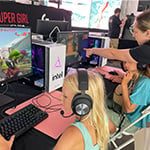 Two young girls are seated at desktop computers playing 'Minecraft' while a woman wearing a Super Girl Gamer Pro shirt assists them.