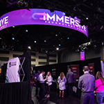 A large room with lots of booths set up, each from a different technology company. People in business-casual clothing are walking around and socializing.