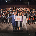 In a large venue full of college graduates, two men in suits stand on opposite sides of a woman in a cap and gown holding a framed photo that reads "Full Sail University - Congratulations to our 100,000th graduate Isabel Miller."