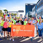 A group of Full Sail faculty, students, and staff posing with a large orange banner that reads “Full Sail University”