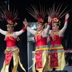 Three women dancing in colorful Indonesian feathered headdresses, yellow skirts, and beaded belts and collars.