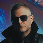 A man in a black leather coat and sunglasses stands before a giant, colorful close-up of an eye glowing with blue and orange light.