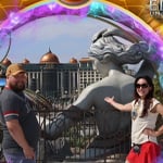A man and woman stand together in front of an attraction featuring a large statue of a woman, a building with a glass dome, and a building with bronze domes.
