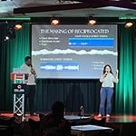 Tyonne Heyward and Ivana Loyola stand onstage in front of an audience. They share a slideshow presentation on the screen behind them.