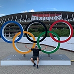 Grad Andrew Molina sits outdoors with his arms spread in front of a set of Olympic rings. There is a stadium with a banner for the 2020 Tokyo Olympics behind him.