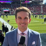 Taylor Schaub stands on the edge of a football field wearing a suit and tie and holding a microphone. Behind him is the football field, stands full of fans, a large screen showing the game, crew on the sidelines, and players on the field.
