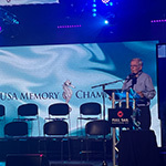 A man wearing glasses and a collared shirt speaks at a podium on a stage. A screen that says USA Memory Championship is behind him.