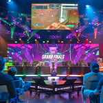 An audience faces a stage where eight competitors sit at consoles. A screen above them shows the game they're playing and another screen displays the NACE Grand Finals 2025 graphic.