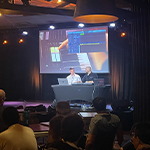 Scott Dickinson and Evan Weiss sit onstage with a keyboard and two laptops. Students sit in an audience facing the stage.