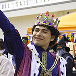 Several students dressed in costumes including a mushroom, a goddess, a viking, and a king stand in a line. Orange tents surround the crowd of students.