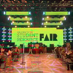 Students and staff sit at tables and interact. A large screen behind them displays “Welcome to the Student Resource Fair” in black text with a green background.