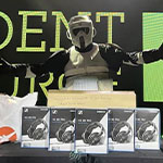 A masked figure stands behind a table piled with boxes of headphones and various technology on stage at the Student Resource Fair.