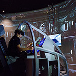 A student wearing a VR headset sits in a space shuttle landing simulator. A large screen displaying a warehouse is in front of them.