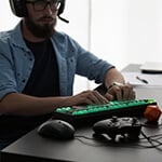 A man wearing a gaming headset sits at a desk with a gaming keyboard, an Xbox controller, and a Dungeons & Dragons die.