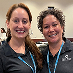 Michelle Moreno and Stacie Aldrich smiling and standing side by side wearing blue lanyards with conference badges and black Full Sail polo shirts.