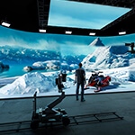 A man wearing jeans, sneakers, and a plaid shirt stands in a production studio. He is facing a large LED screen showing an icy mountain range and a snowmobile.