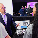 Man wearing glasses and a blue suit speaks to guests at Career Fair while man in back tries on Virtual Reality goggles.