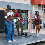 Three performers stand onstage. Two are holding microphones and singing, and one is playing an electric guitar.