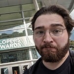 Ryan Miller pictured from above the shoulders in a black leather jacket with silver wireframe glasses in front of a sign reading “Game Developers Choice Awards.”