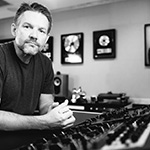 Brad Blackwood sits at an audio console in a studio with framed gold records on the wall behind him.
