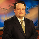 Dan Hanson sits at a news desk in a television studio. He is smiling and wearing a black suit with a yellow tie.