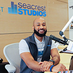 Full Sail grad Anthony Grier sits in front of an audio board at Orlando Health Arnold Palmer Hospital for Children’s Seacrest Studios, smiling.