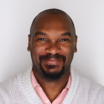 Quentin stands in front of a white wall wearing a white cardigan over a pink polo. His arms are crossed and he is smiling.