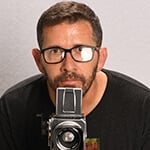 Sebastian Krys crouches behind a video camera. He is wearing black-rimmed eyeglasses and a black t-shirt with a logo on it.