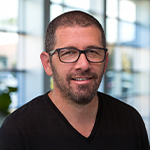 Sebastian Krys smiles at the camera in a lobby with large glass windows. He is wearing glasses and a black t-shirt.