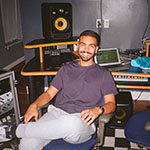 A smiling man is seated casually in a music studio, surrounded by audio equipment, a keyboard, speakers, and a laptop on a desk.