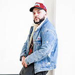 A man wearing a red baseball cap and a denim jacket is pictured leaning against a concrete pillar in a brightly lit room.