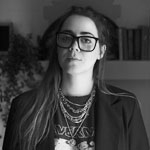 Veronica Concha stands in front of a bookshelf in her home. She is wearing a dark blazer, a band t-shirt, silver necklaces, and glasses.