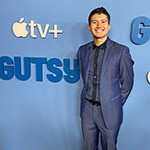 A person with short, dark hair is smiling while wearing a blue suit against a light blue step and repeat that shows the logos for ‘Gutsy’ and Apple TV+.