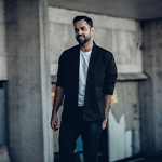 A well-dressed man in a white t-shirt and black blazer posing in front of a worn-down building.