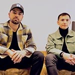 Two men seated side by side on a light brown leather couch against a white backdrop.