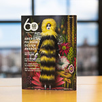 Graphic Design USA’s April 2023 issue sits upright on a wooden table. The cover features a female doll wearing a long furry black-and-yellow dress with a hood. She is standing in front of a psychedelic floral background.