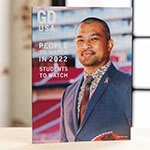 A Graphic Design USA magazine stands upright on a desk in a backlit room. On the cover, a man wearing a blue suit with a pink ribbon pin and holding an NFL football smiles for the camera. The cover reads, People to Watch in 2022, Students to Watch.