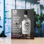 A copy of 'Graphic Design USA' magazine sits atop a white surface against a blurred background. The magazine cover depicts a bottle of gin and a martini glass against a gray background and reads '61st Anniversary American Package Design Awards.