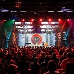 A person wearing a suit speaks on a stage in front of a large crowd. There is a large screen with a logo that says Hall of Fame XII behind them. Red, blue, and white lights illuminate the room.