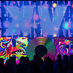 A large sculpture of the letters “HOF” sits atop the Full Sail Live Venue stage with different multicolored images projected onto each letter. The letters are in front of a large purple screen featuring the Roman numeral “XV” in large font. In front of the stage is a black silhouette of the crowd.