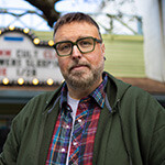 Aaron B. Koontz stands in front of a movie theater. He is wearing green glasses, a green sweatshirt, and a collared plaid shirt.