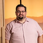 A man with glasses and a beard in a pink button-down stands next to a wooden panel, with a warm, mountainous sunset background behind him.