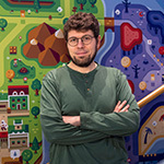 A bearded man in glasses and a green shirt stands with his arms crossed in front of a colorful, game-inspired illustrated mural.