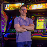 Jonathan Mayer smiles and stands in front of a row of arcade game machines for ‘Tron’, ‘Kung-Fu Master’, ‘Dig Dug’, and ‘Donkey Kong’.
