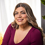 Katrina Mena Rick smiles at the camera. She is sitting on a gray couch in a living room and wearing a maroon V-neck top.